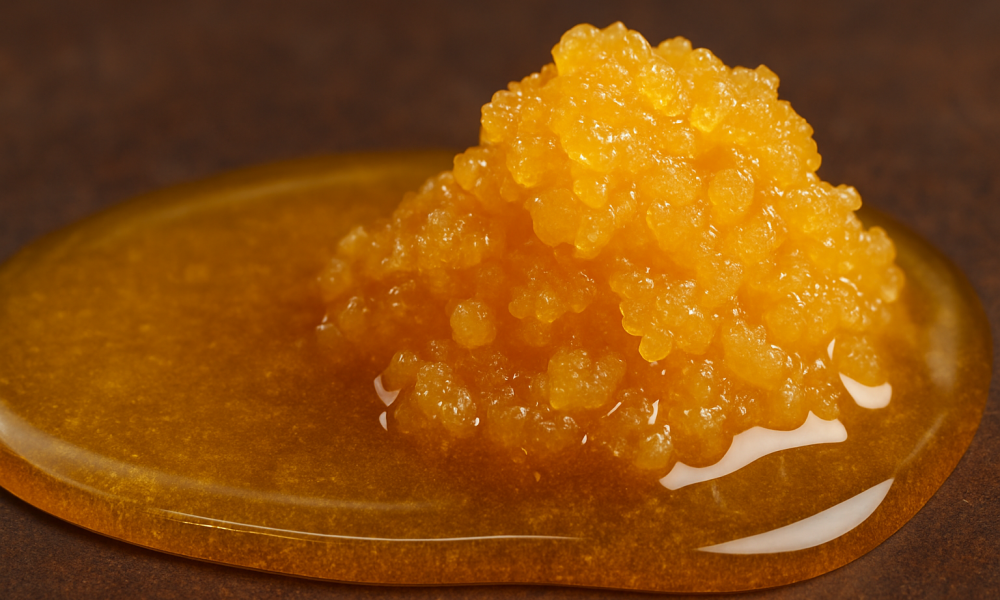 Live resin vs. Distillate of Cannabis (2) A horizontal close-up digital photograph shows a golden, glossy mound of live resin on a smooth surface. A stream of translucent cannabis distillate is being poured next to it, forming a shiny, syrup-like pool around the base of the live resin. The contrast in texture and opacity between the thick, crystal-like live resin and the clear, liquid distillate visually highlights their differences.